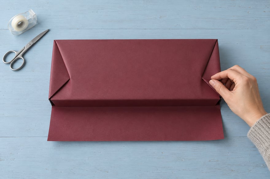 An overhead view of a hand creasing burgundy wrapping paper around a flat box on a pale blue table